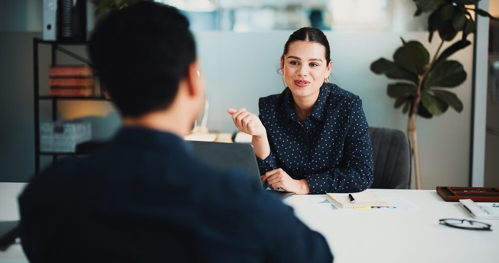 Manager speaking with employee across a desk, highlighting personal communication during mergers and acquisitions to employees.