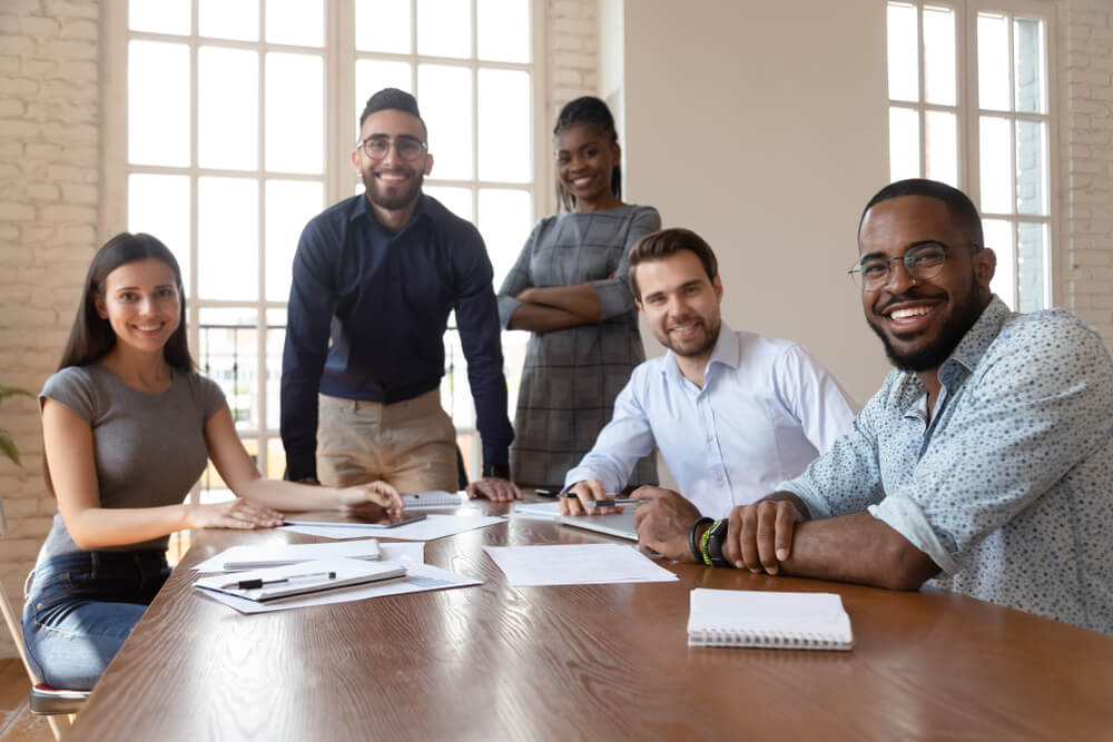 Portrait of happy diverse young businesspeople gather at boardroom table brainstorming discussing business ideas together, multiethnic millennial colleagues look at camera posing at meeting.