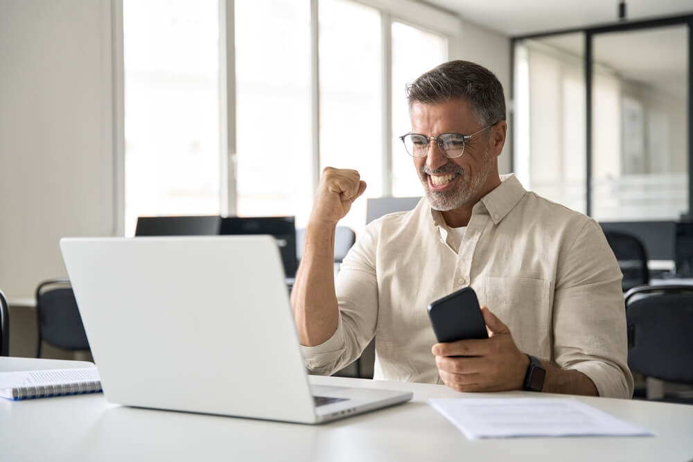 Smiling entrepreneur pumps his fist while checking a laptop and smartphone, illustrating how do franchise owners get paid?
