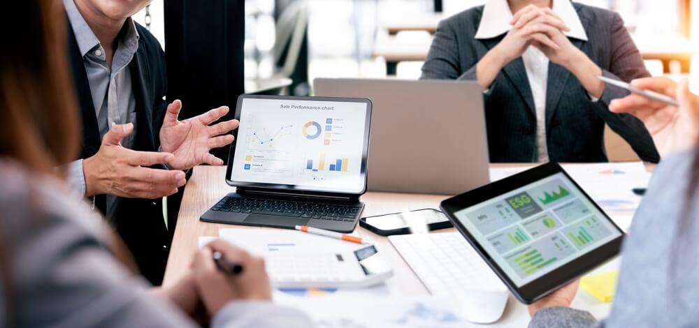 Business professionals meet around a table with a laptop and tablet displaying charts and dashboards, discussing insurance industry trends.